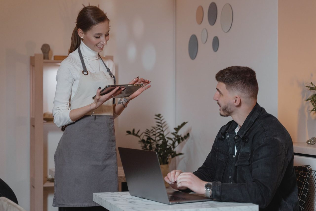 A smiling waitress takes an order on a tablet in a cosy café